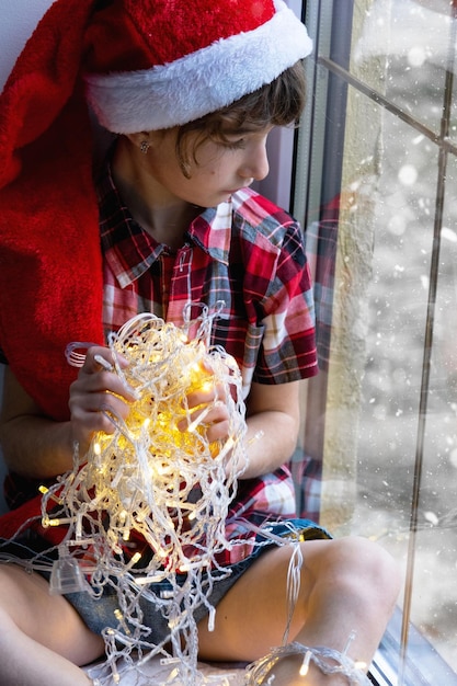 Uma garota segura uma guirlanda em suas mãos sentada no parapeito da janela e no clima festivo do Natal no ano novo