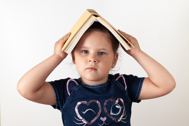 Uma garota séria em um vestido jeans segura um livro na cabeça, uma criança, isolada.