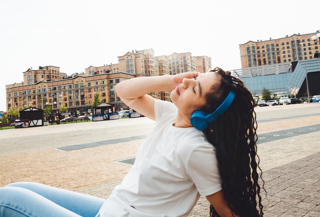Uma garota sorridente com dreadlocks em fones de ouvido está sentada no chão no parque Uma jovem feliz relaxando com fones de ouvido curtindo música Espaço para copiar a geração z