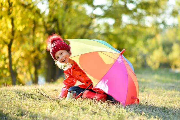 Uma garota sorridente com um chapéu vermelho e uma jaqueta caminha com um guarda-chuva de arco-íris na natureza Atmosfera de outono