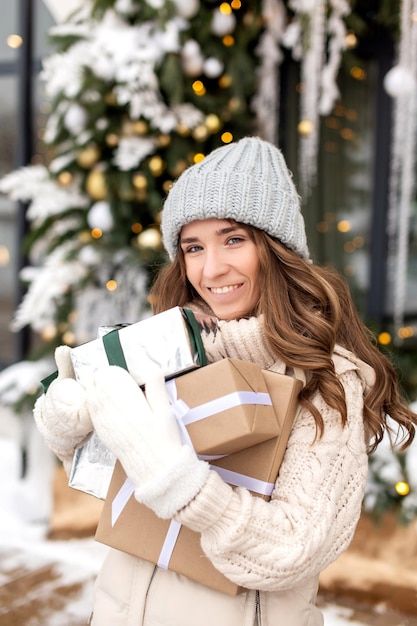 Uma garota sorridente de chapéu abraça caixas de presentes na rua no inverno perto de lojas