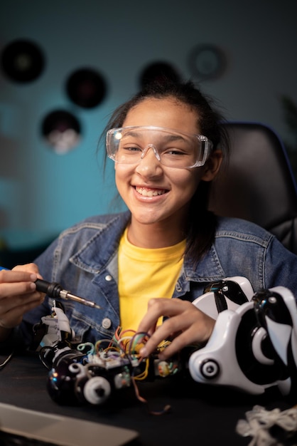 Uma garota talentosa sorridente mexe em seu quarto em um robô que ela usa óculos de segurança usa uma solda