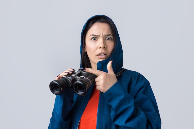 Uma garota turista com uma capa de chuva azul segura binóculos nas mãos e olha para longe, espiões.