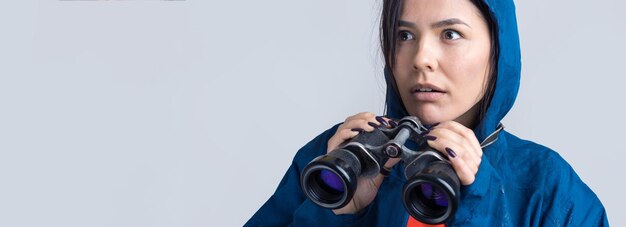 Foto uma garota turista em uma capa de chuva azul segura binóculos nas mãos e olha para os espiões à distância