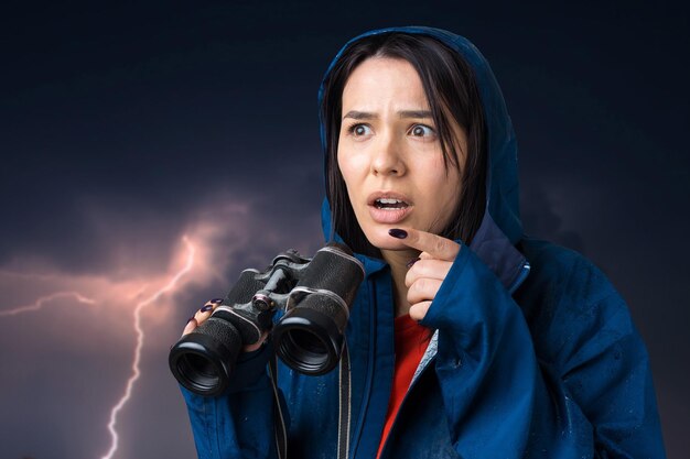 Uma garota turista em uma capa de chuva azul segura binóculos nas mãos e olha para os espiões à distância