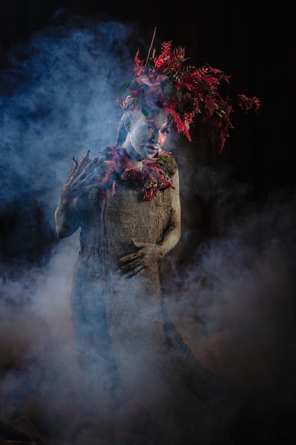 Foto uma garota untada com argila em um vestido cimentado. a modelo tem um toucado feito de flores. fumaça por trás