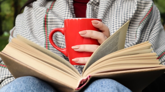 Uma garota vestida com um casaco e um lenço na floresta de outono segura um livro e um copo com uma bebida quente em seu close-up de mãos em um parque da cidade em um dia quente. O conceito de leitura, relaxamento e conforto.