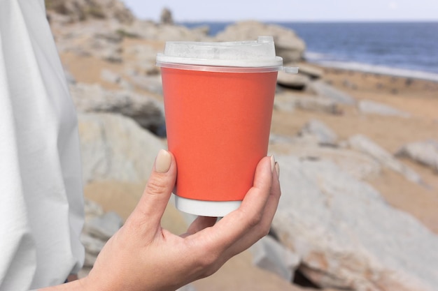 Foto uma garota vestindo uma camiseta branca e jeans azul está segurando uma caneca vermelha para café para viagem contra o pano de fundo de uma praia do mar com areia e pedras cinzentas