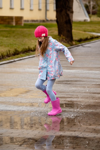 Uma garotinha bonitinha em uma capa azul botas rosa e um chapéu rosa atravessa poças e se diverte