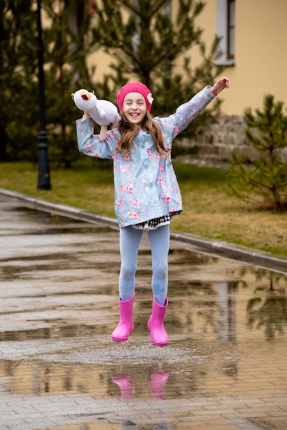Uma garotinha bonitinha em uma capa azul botas rosa e um chapéu rosa atravessa poças e se diverte