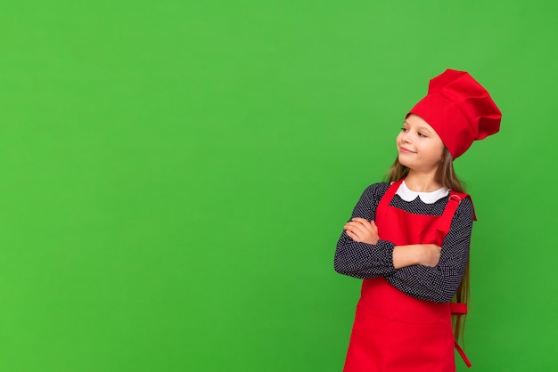 Uma garotinha chef vestida com um avental e um boné de cozinha cruzou os braços no peito e olha para o seu anúncio em um fundo verde isolado