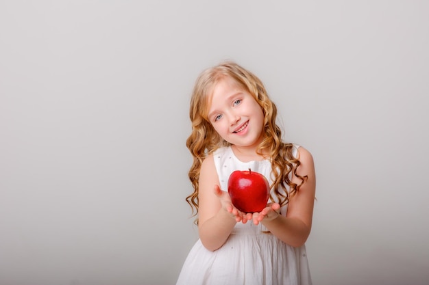 Uma garotinha com cabelo comprido e um vestido branco segura uma maçã em um fundo branco