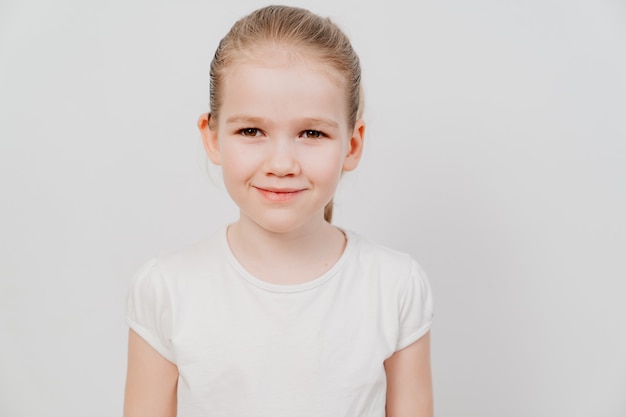 Uma garotinha com cabelos presos em uma camiseta branca fica em uma superfície branca e sorri.