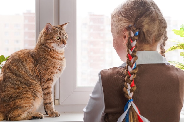 Uma garotinha com fitas no cabelo e um gato se olham em pé na janela