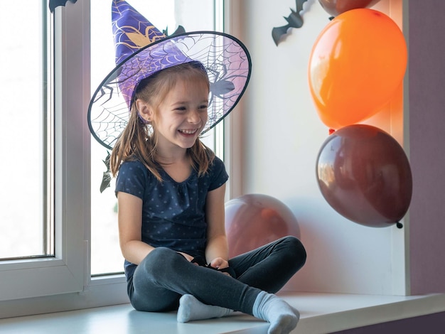 Uma garotinha com um chapéu de bruxa está sentada no peitoril da janela sorrindo olhando para longe feliz Halloween