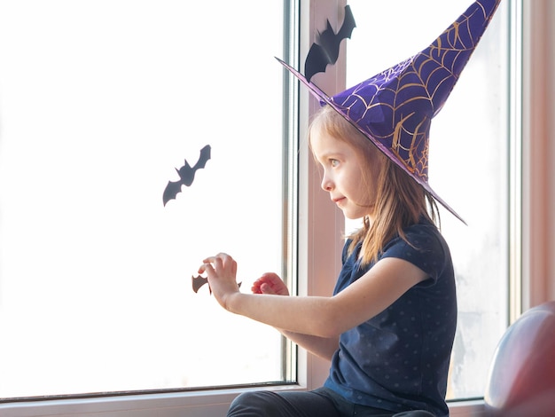 Uma garotinha com um chapéu de bruxa olha pela janela no feliz dia das bruxas