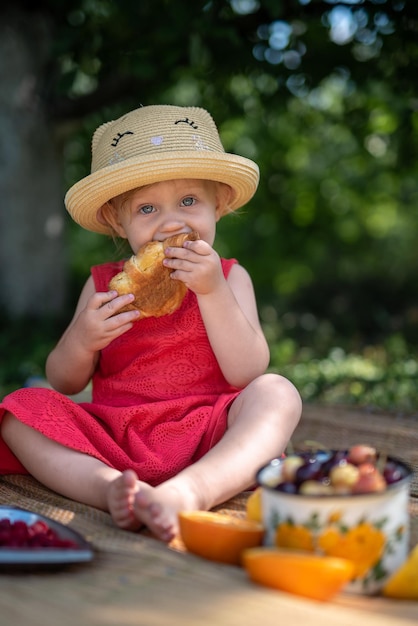 Uma garotinha com um chapéu de palha e um vestido vermelho com os pés descalços come um croissant