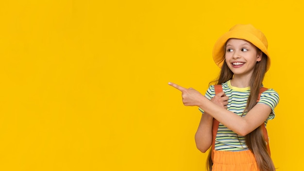 Uma garotinha com um chapéu de verão e uma mochila aponta o dedo para o seu anúncio A criança está indo para um acampamento Amarelo isolado fundo brilhante Copiar espaço Banner