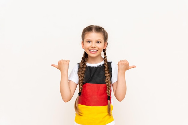 Uma garotinha com uma bandeira alemã em uma camiseta com cabelo comprido em tranças aponta para cima com o dedo indicador de lado para seu anúncio em um fundo branco isolado Cursos de idiomas