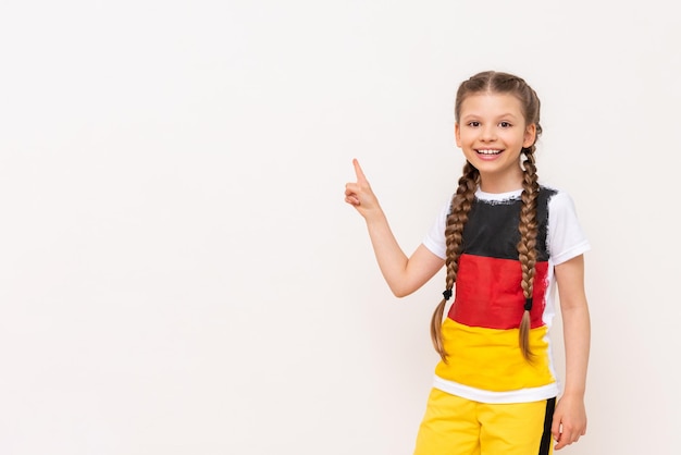 Uma garotinha com uma bandeira alemã em uma camiseta com cabelos longos em tranças aponta para cima com o dedo indicador de lado para seu anúncio em um fundo branco isolado Cursos de idiomas Copiar espaço