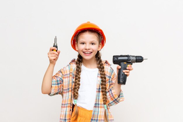 Uma garotinha com uma chave de fenda elétrica em um capacete protetor laranja sorri amplamente em um fundo branco isolado O conceito de renovação no quarto de uma adolescente