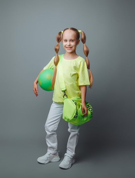 Uma garotinha com uma espada verde na mão sorri para a câmera isolada em um fundo cinza um pequeno atleta com uma bola esportes infantis