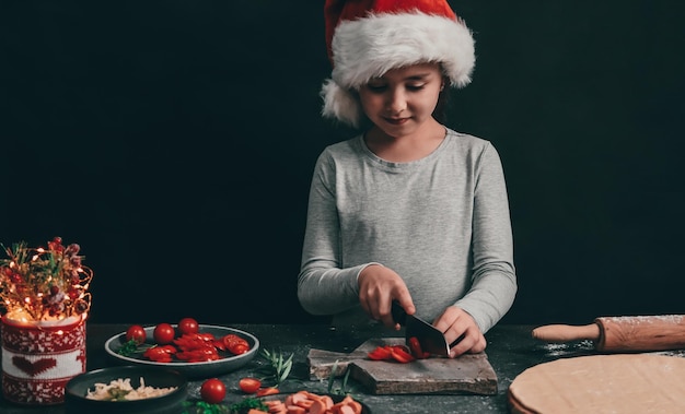 Uma garotinha corta com uma grande faca preta tomate cereja em uma placa de madeira cinza