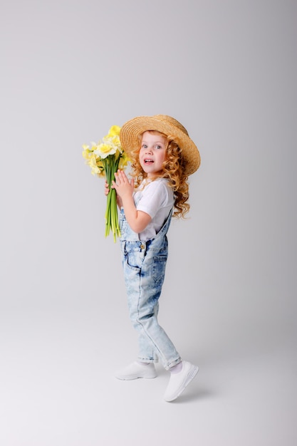 uma garotinha de cabelos encaracolados com um chapéu de palha segura um buquê de flores de narciso amarelo da primavera em um whi