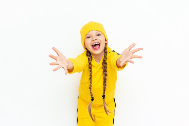 Uma garotinha de chapéu e agasalho amarelo estica os braços para a frente e sorri amplamente contra um fundo branco isolado