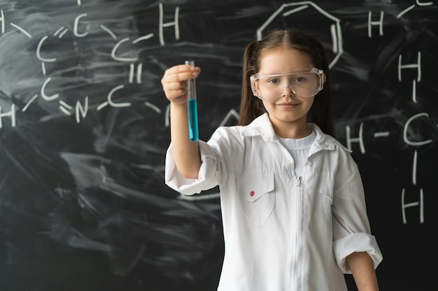 Uma garotinha de colegial de óculos e jaleco branco está perto do teste da aula de química no quadro-negro ...