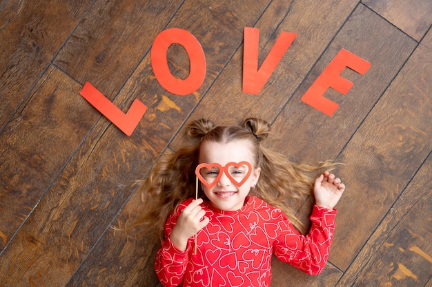 Uma garotinha de pijama vermelho com a inscrição amor encontra-se em um fundo de madeira marrom escuro no chão e ri segurando óculos em forma de coração o conceito de dia dos namorados