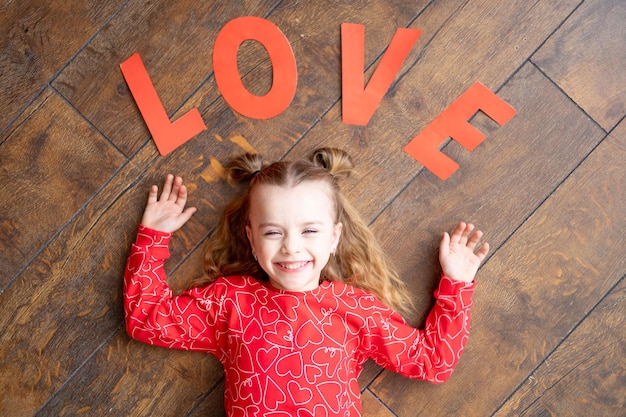 Uma garotinha de pijama vermelho com a inscrição Love encontra-se em um fundo de piso de madeira marrom escuro e ri do conceito de dia dos namorados um lugar para texto