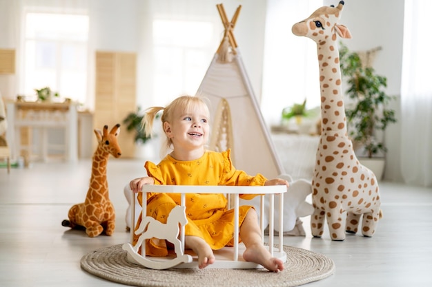 Uma garotinha de vestido amarelo brinca com uma girafa em casa com uma tenda em uma sala iluminada e sorri uma infância feliz