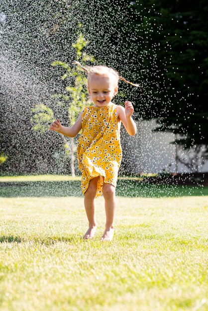 Foto uma garotinha de vestido amarelo corre por um aspersor de água.