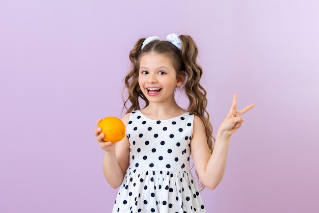 Uma garotinha de vestido de bolinhas segura uma laranja e mostra um coelho Vitaminas para crianças