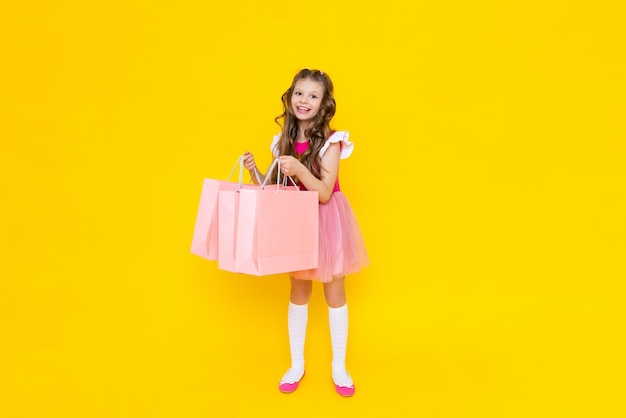 Uma garotinha de vestido rosa segura grandes sacolas de compras em um fundo amarelo isolado compras de primavera na loja grandes promoções de mercadorias