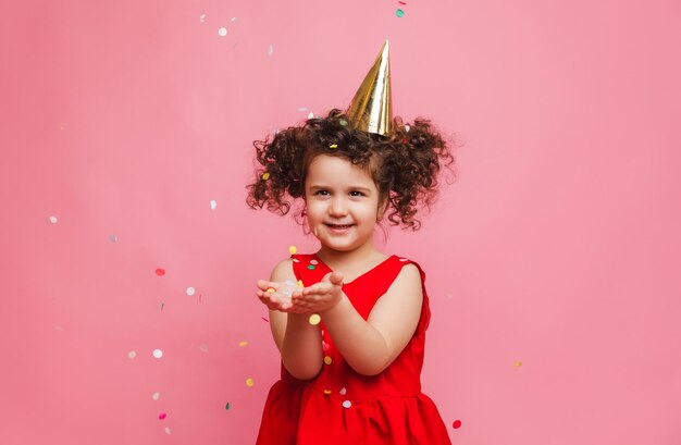 Foto uma garotinha de vestido vermelho comemora seu aniversário soprando e pegando confete em um fundo rosa