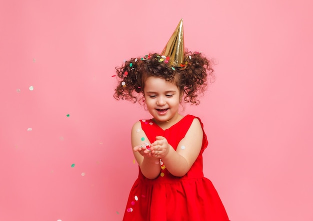 Uma garotinha de vestido vermelho comemora seu aniversário soprando e pegando confete em um fundo rosa