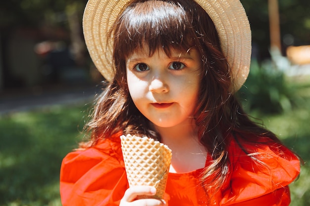 Uma garotinha de vestido vermelho e chapéu de palha come sorvete em um cone no parque