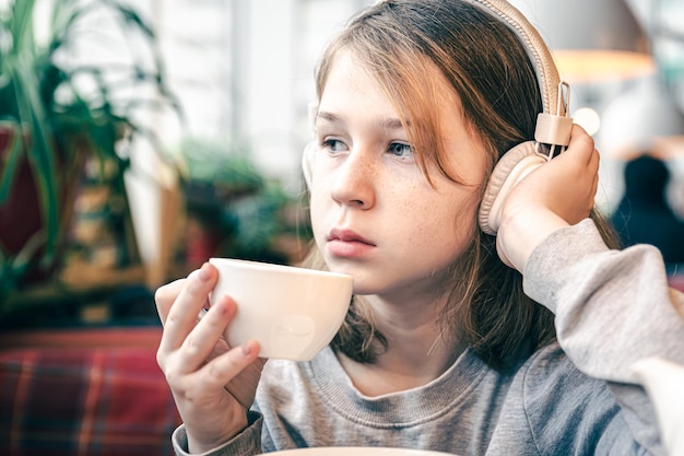 Uma garotinha em fones de ouvido em um café com uma xícara de chá