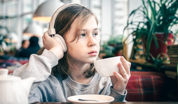 Uma garotinha em fones de ouvido em um café com uma xícara de chá