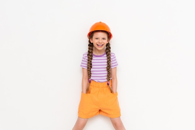 Uma garotinha em um capacete de construção e tranças segura as mãos nos bolsos em um fundo branco isolado A profissão de engenheiro para uma criança