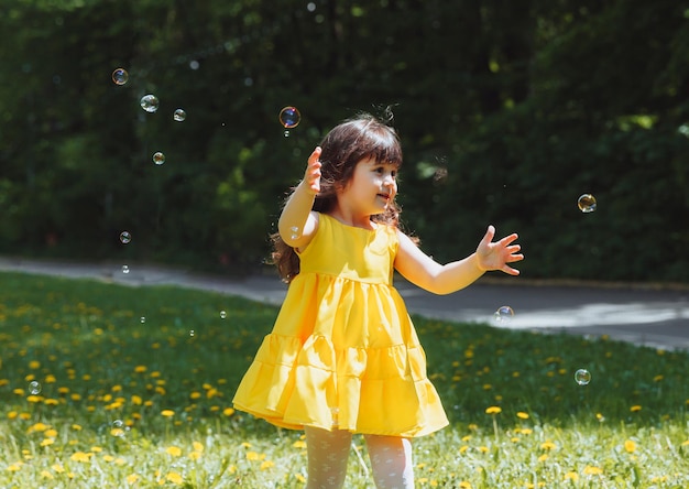 Uma garotinha em um vestido amarelo de verão pega bolhas de sabão na grama no parque coroa de dentes de leão