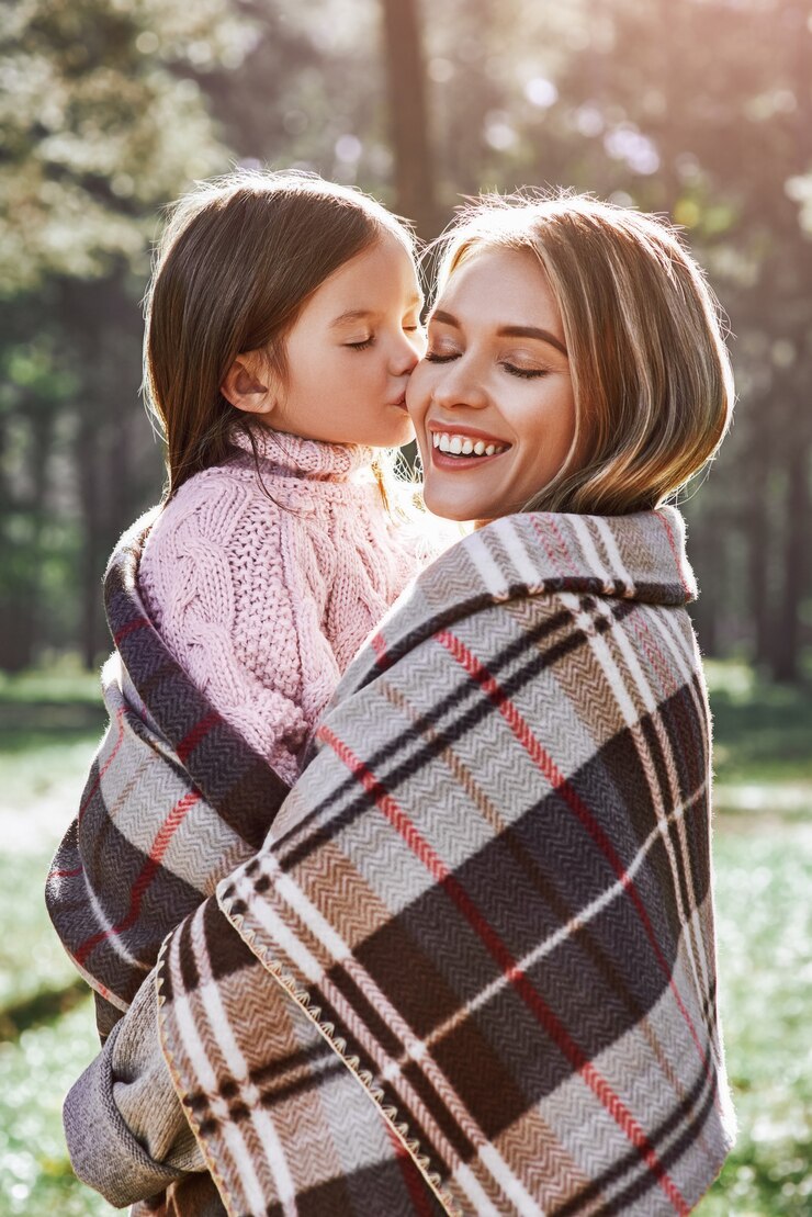Uma garotinha encaracolada de suéter está beijando a mãe na floresta. Ela está vestindo um suéter rosa e uma jovem está usando um suéter cinza. Eles estão embrulhados em um cobertor