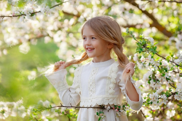 uma garotinha engraçada e fofa em um vestido na primavera perto de uma árvore florida está rindo fotografia gentil