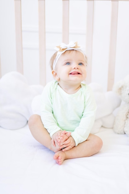Uma garotinha engraçada e fofa mostra a língua em um berço em uma cama de algodão branco em uma roupa verde em um quarto infantil brilhante e sorri