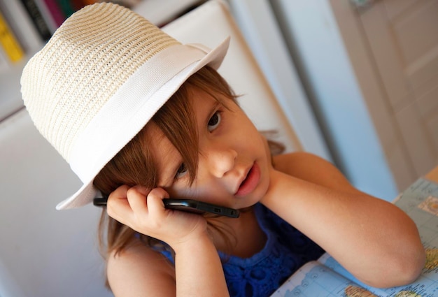 Uma garotinha entediada falando com celular retrato da menina bonitinha em roupas casuais de verão e chapéu branco com smartphone conceito de infância e tecnologia tédio aborrecimento em crianças em casa