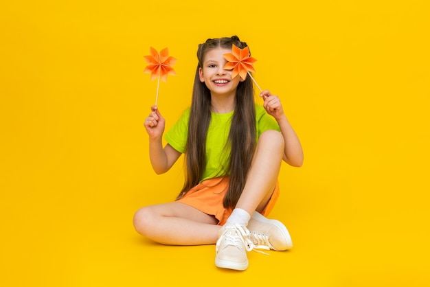 Uma garotinha está brincando com flores de papel em palitos Um moinho de origami Criança feliz segura artesanato em papel Criatividade infantil Uma linda criança está sentada em um fundo amarelo isolado