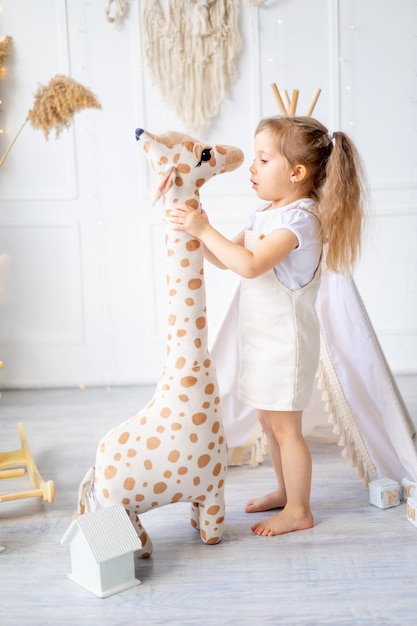 Uma garotinha está brincando com uma grande girafa tão alta quanto ela está em casa em um berçário brilhante e rindo