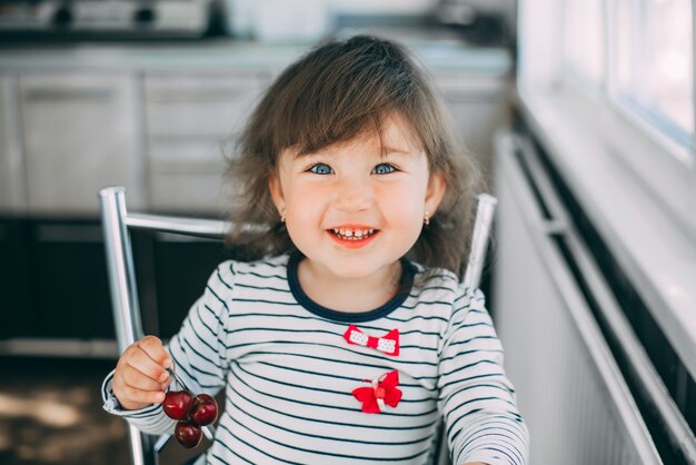 Uma garotinha está comendo cerejas, segurando frutas nas mãos e sorrindo fofamente